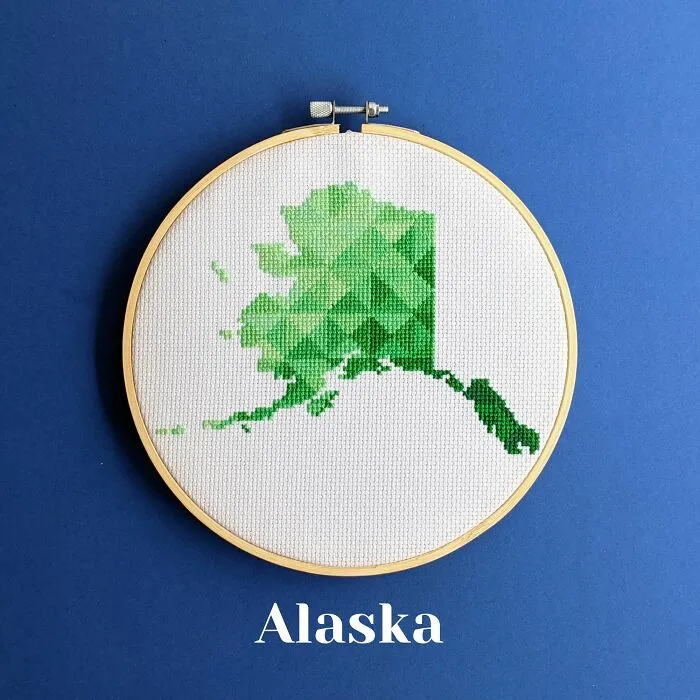 Cross-stitched map of Alaska in green shades displayed in an embroidery hoop on a blue background.