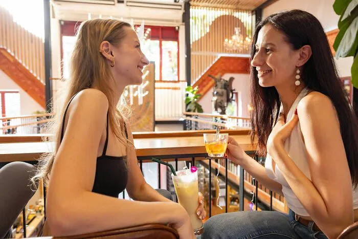 Two women smiling and enjoying drinks at a bar during dates that went sour, using the bar’s safe word.