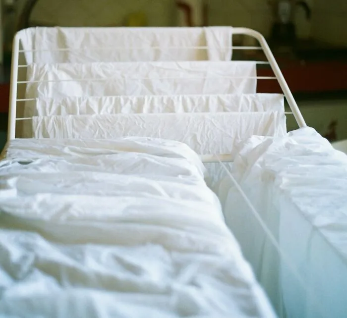 A white hospital bed with crumpled sheets and a drying rack holding white linens in a softly lit room.