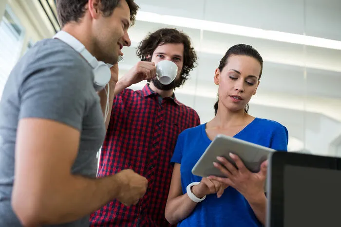 Three corporate colleagues in a casual office setting discussing work while one looks at a tablet, illustrating calling out lies.