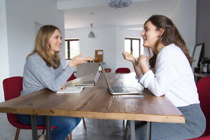 Two women in a corporate setting talking and smiling across a wooden table with laptops, illustrating calling out corporate pals’ lies.