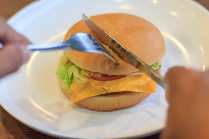 Person cutting a cheeseburger with knife and fork, illustrating things non-Americans do like breaking spaghetti for Italians.