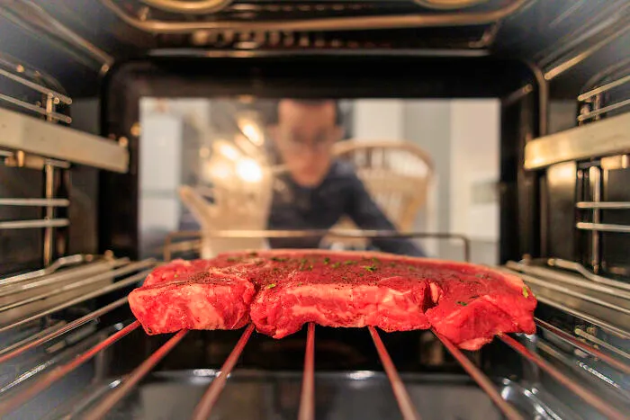 Raw steak inside an oven with a person blurred in the background, illustrating Americans reveal non-Americans food habits.