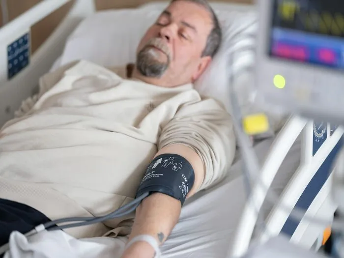 Elderly man resting in hospital bed with blood pressure cuff, showing a typical hospital experience in a medical drama setting.