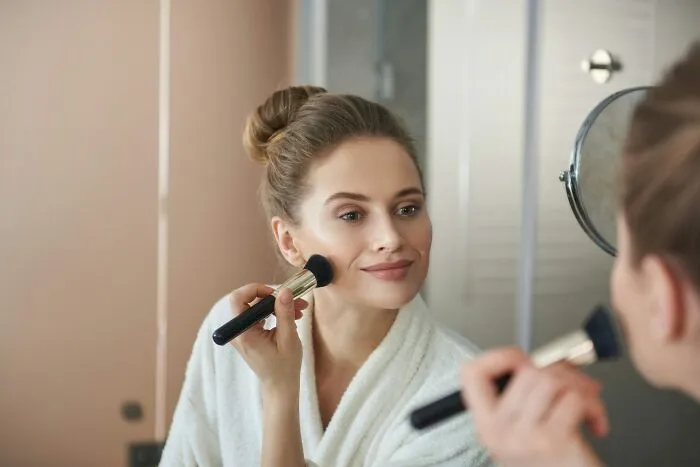 Young woman in a white robe applying makeup with a brush, reflecting calm despite no peaceful solution to our current situation.