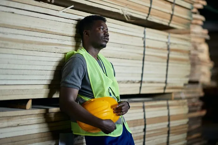 Man wearing safety vest and holding yellow helmet, leaning against stacked wooden planks reflecting current situation challenges.