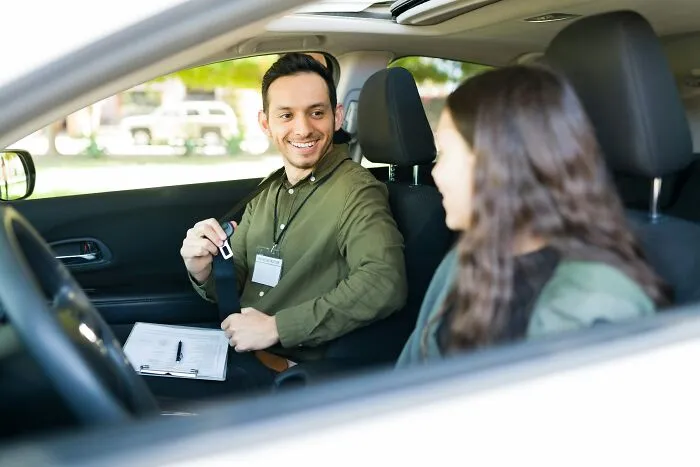 Man instructor helping woman with seatbelt during a driving lesson inside a parked car outdoors.