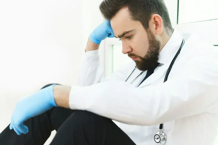Young male doctor wearing gloves and a stethoscope, sitting stressed in a hospital during an intense medical drama.