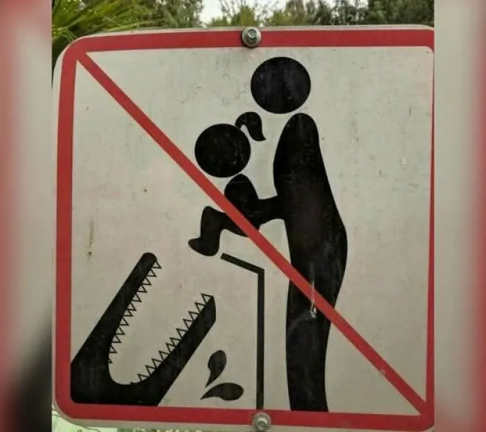 Warning sign showing a person holding a child near a crocodile with a red line, a hilarious sign adding chaos to the world.