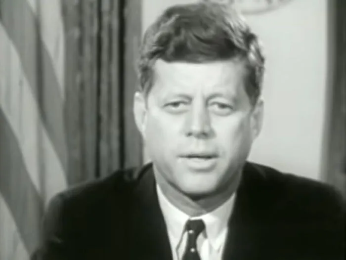 Black and white portrait of a historical figure speaking in a formal setting with a flag in the background.