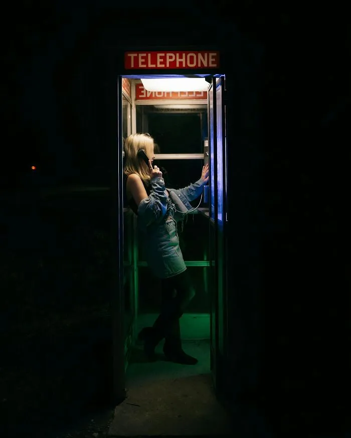 Young woman using a telephone booth at night, highlighting childhood joys lost with smartphones in a dark setting.