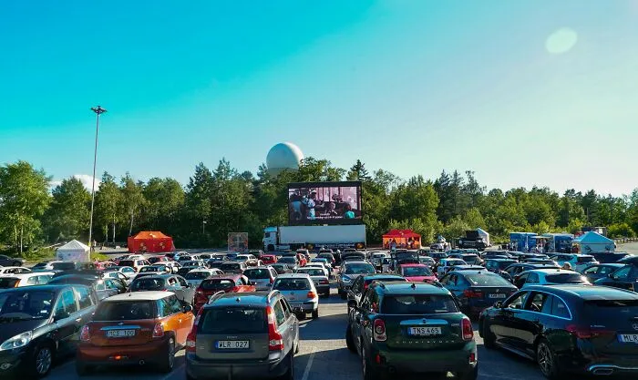 Cars parked at an outdoor drive-in movie theater under a clear blue sky with trees in the background.