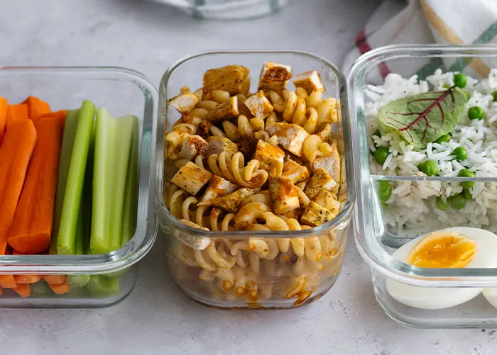 Meal prep containers with carrot sticks, celery, pasta with chicken, and rice with peas and a boiled egg, healthy foods.