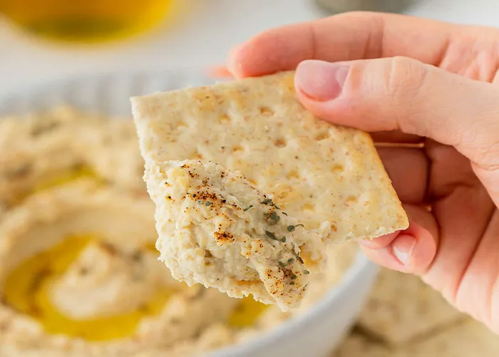 Hand holding cracker with dip, illustrating healthy foods you might want to skip eating according to dentists and doctors.