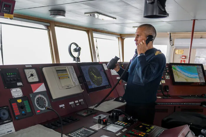 Man in a navy uniform using a radio on a ship’s bridge surrounded by navigation and communication equipment, representing professions.
