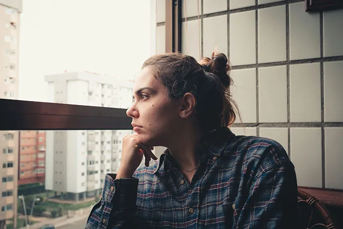 A young woman in a plaid shirt looking out the window with a thoughtful expression, reflecting on family and relationships.