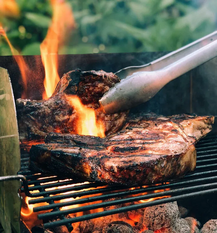 Grilled meat over open flame with tongs turning food, highlighting tobacco and health risks related to smoking.