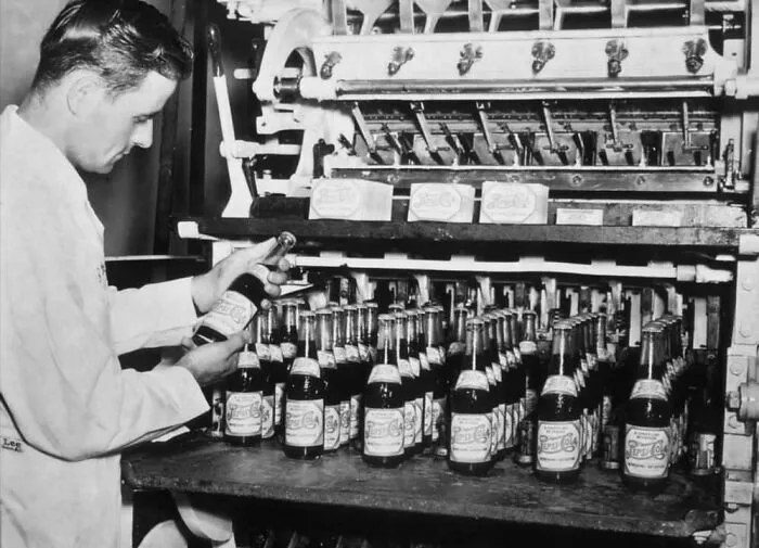 Hombre inspeccionando botellas en línea de producción antigua, mostrando un lado distinto de la historia industrial.