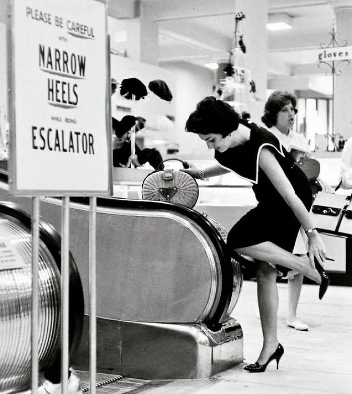 Mujer con tacones altos ajustándose el zapato junto a una escalera mecánica, foto histórica mostrando un lado distinto de la historia.