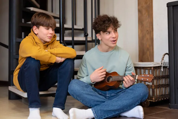 Two teenage boys sharing raw and emotional confessions while sitting casually, one playing a small guitar indoors.