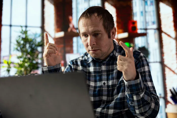 Man in a plaid shirt sharing raw and emotional confessions while working on a laptop in a sunlit room.