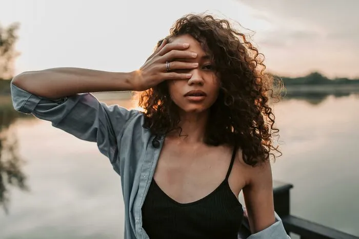 Young woman with curly hair by the lake covering one eye, reflecting raw and emotional confessions and heavy secrets.