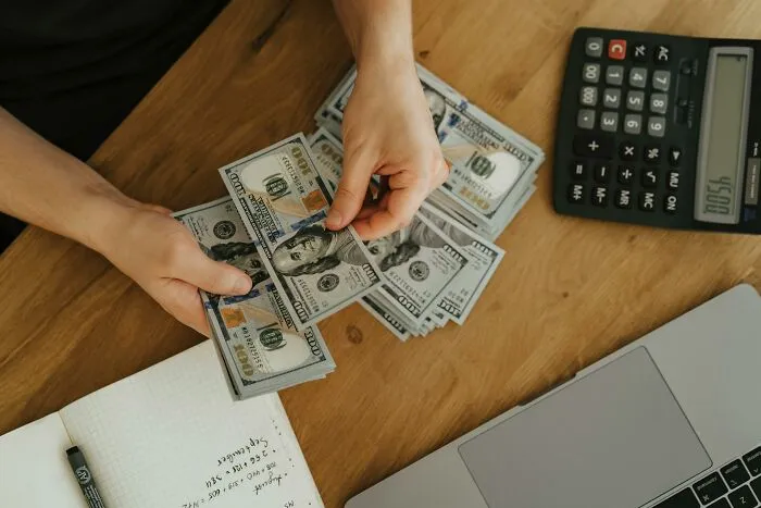 Hands counting hundred-dollar bills on wooden desk with calculator laptop and notebook showing heavy secrets concept.