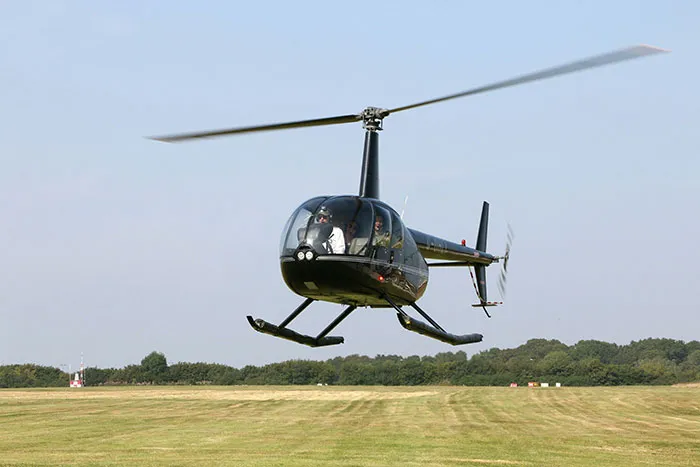 Black helicopter flying low over open grassy field with trees in the background during clear day.