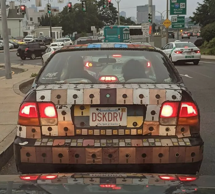Car with outrageous custom metal plate mods covering rear, showcasing extreme and unique car modification creativity on city street.