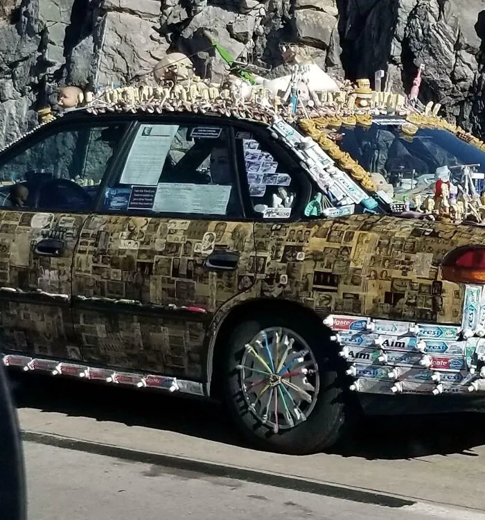 Car covered with photos, dolls, dental products, and various items, showcasing outrageous car mods on a city street.