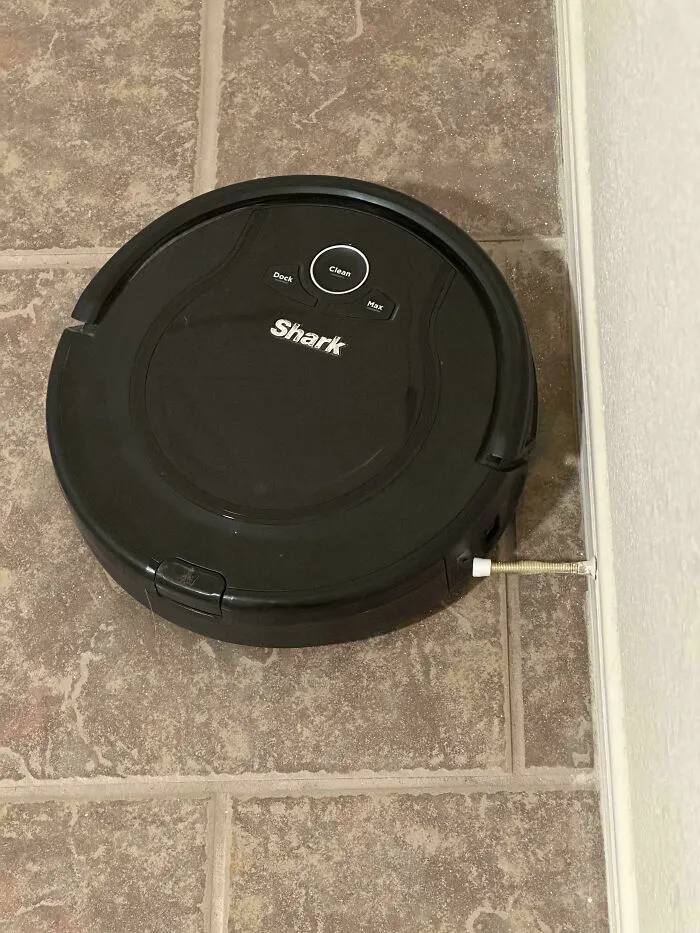 Black Shark robot vacuum stuck on a tile floor with a spring jammed between the cleaning device and the wall, mildly interesting scene.