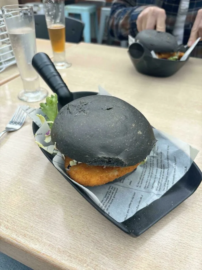 Black bun fish sandwich served in a frying pan on printed paper, an unusual meal presentation style.