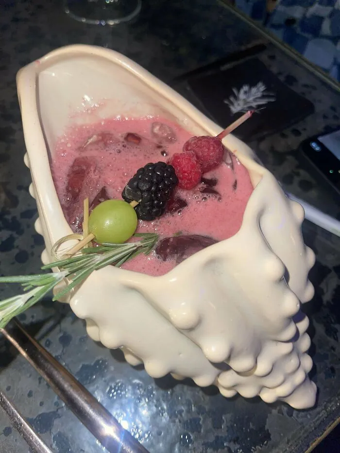 Unholy meal served in a shell-shaped container with pink drink and fruit skewer including grape and berries.