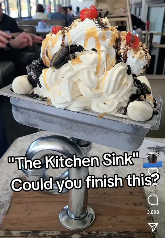 Unholy meal served as a dessert overflowing with whipped cream, cherries, cookies, and syrup in a metal kitchen sink bowl.