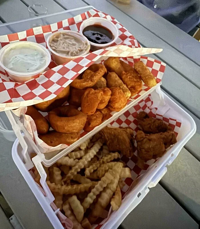 Two-tiered takeout container with fried food, crinkle fries, and dipping sauces served in unholy meal presentation.