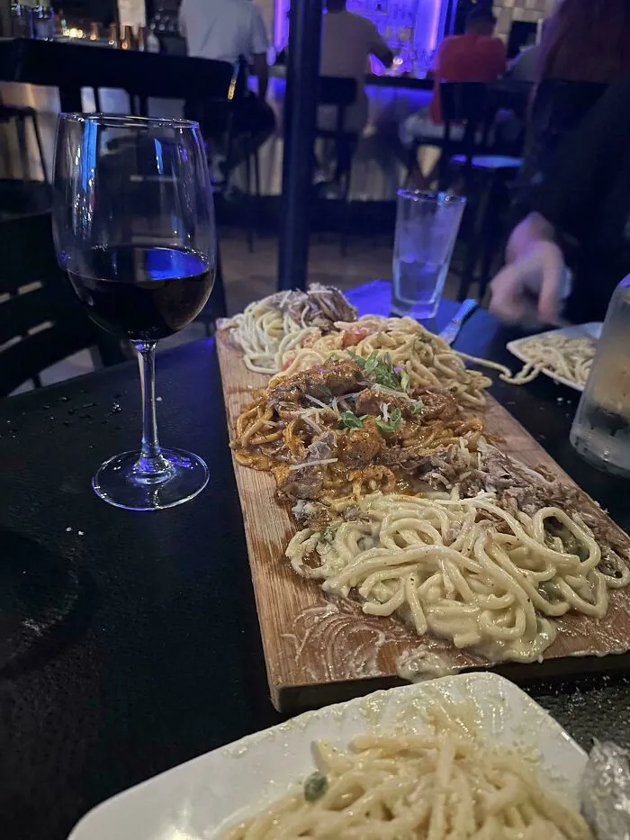 Multiple pasta dishes served untraditionally on a wooden board alongside a glass of red wine in a dim restaurant setting.