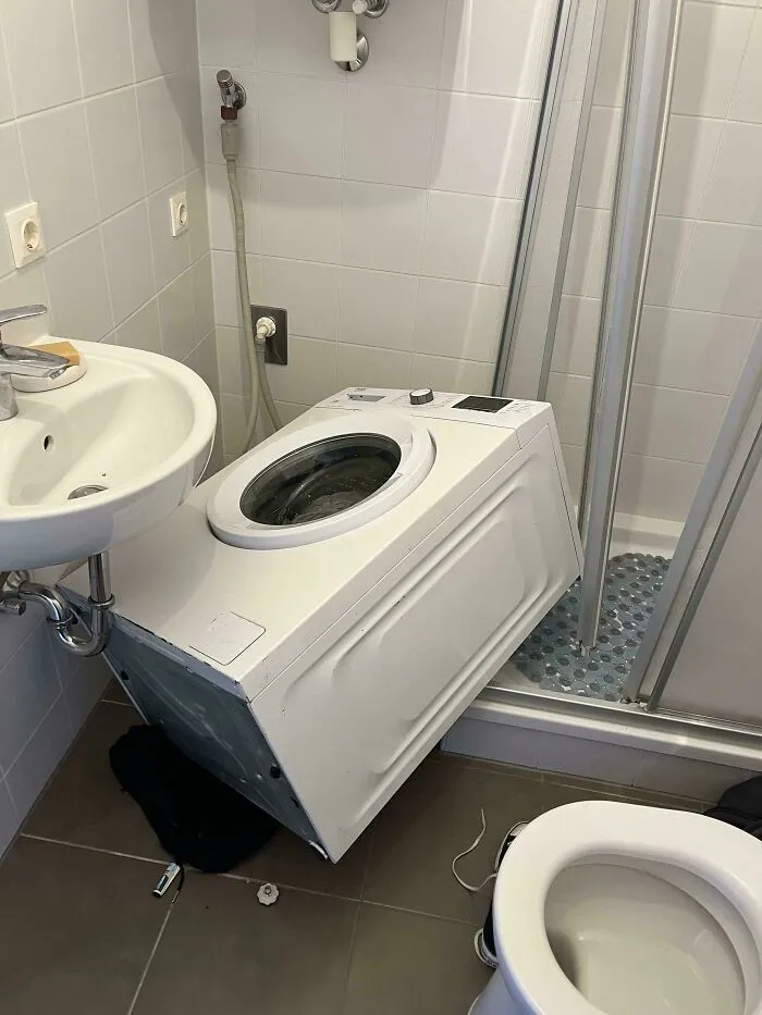 Washing machine fallen over in a small bathroom corner causing a funny unfortunate household mishap.