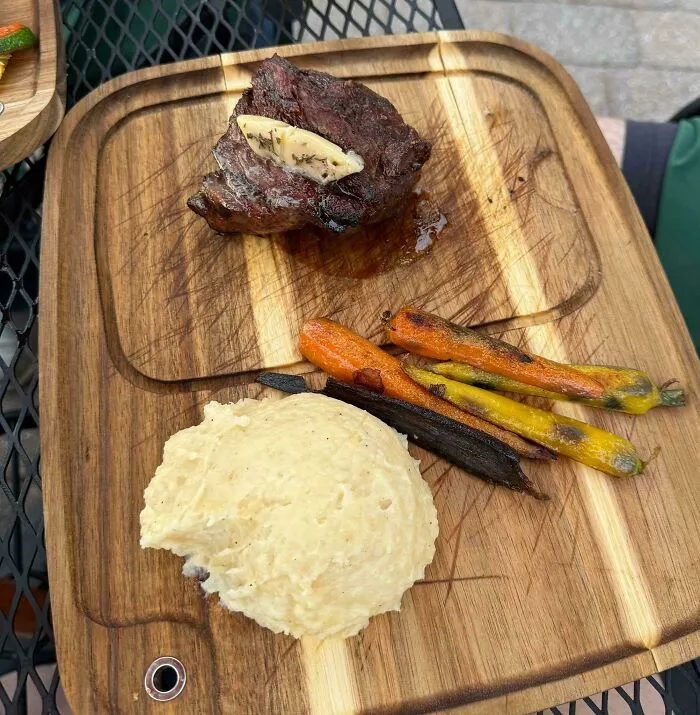 Steak with herb butter, grilled carrots, and mashed potatoes served on a wooden board in an unusual meal presentation.