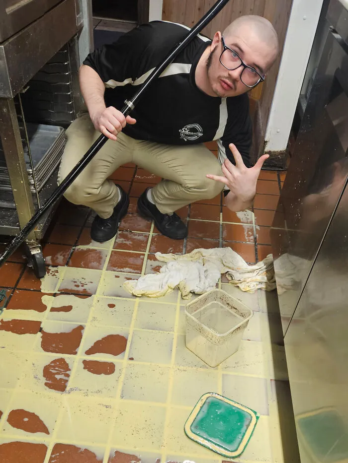 Hombre con gafas y camiseta negra limpiando un gran desastre en el suelo en una situación de día peor en el trabajo.