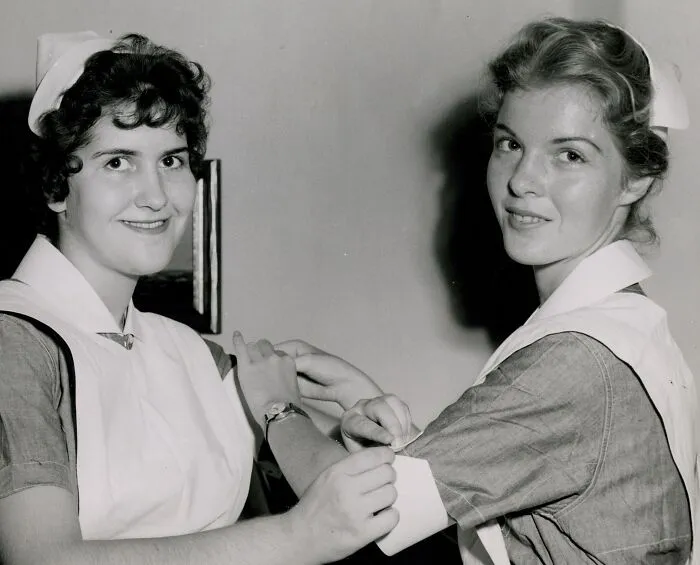 Two vintage nurses smiling while preparing an arm bandage, showcasing classic nursing uniforms and care.