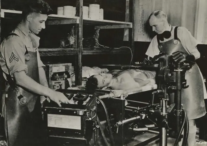 Two men operate unsettling medical devices on a patient lying on a table in a vintage medical setting.