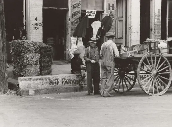 Black and white Great Depression photo showing men with a horse-drawn wagon outside a storefront with hay bales nearby.