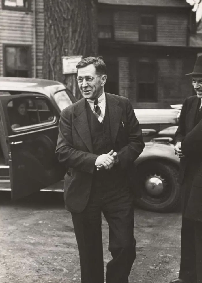 Man in suit and glasses standing next to a car during the Great Depression, captured in a historic black and white photo.