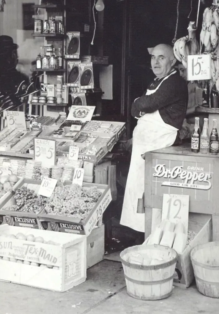 Shopkeeper in apron standing at a fruit and grocery stand during the Great Depression era in a black and white photo.