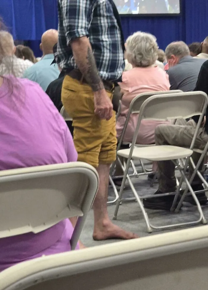Man standing barefoot in a crowded school meeting room with folding chairs and people seated around him.
