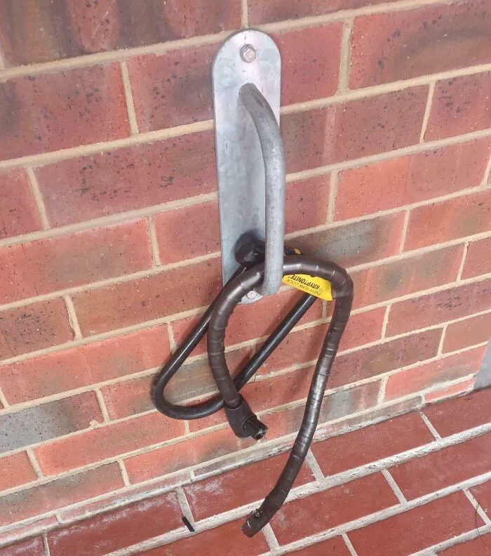 Broken and inadequate bike lock hanging on a metal hook on a brick school wall, showing poor school facility quality.