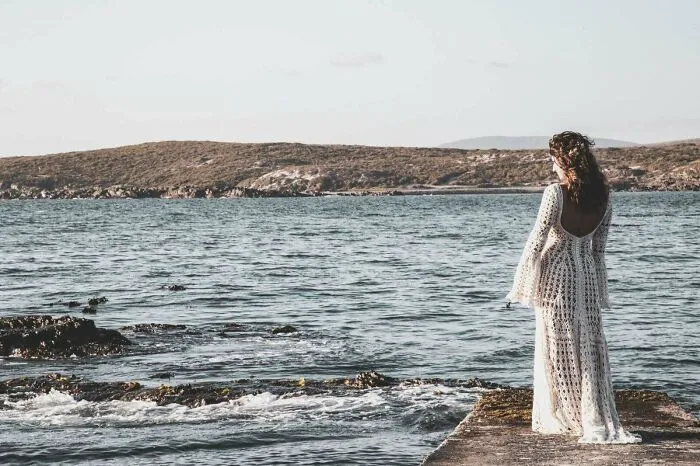 Woman wearing a crocheted dress standing on a pier overlooking the sea with hills in the background, showcasing crochet craftsmanship.