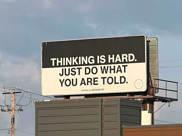 Billboard with text criticizing modern life mindset, illustrating boring dystopia in contemporary urban environment.