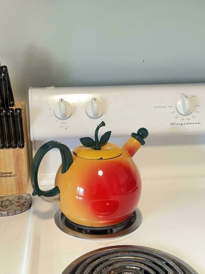 Colorful apple-shaped teapot on stove burner among thrift store finds displayed in a kitchen setting.