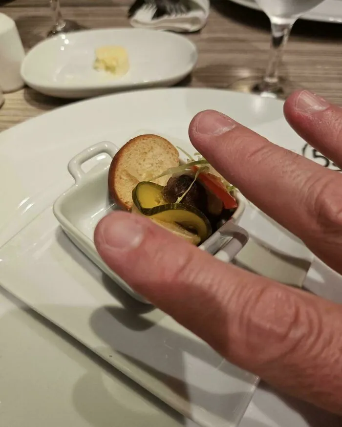 Tiny appetizer dish with pickles, bread, and vegetables served in a small white bowl at a restaurant table.